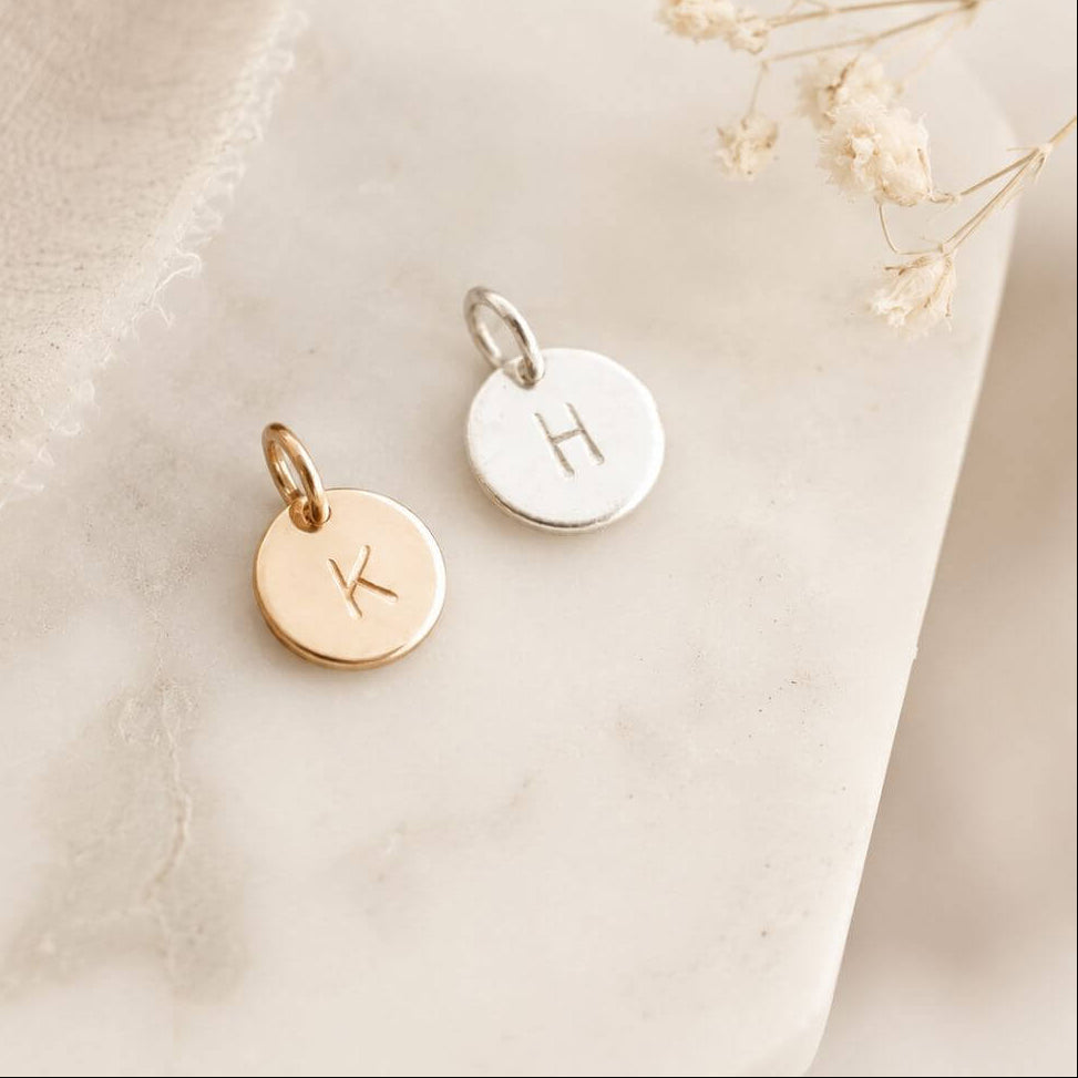 Two gold and silver pendants with initials on a soft beige fabric background